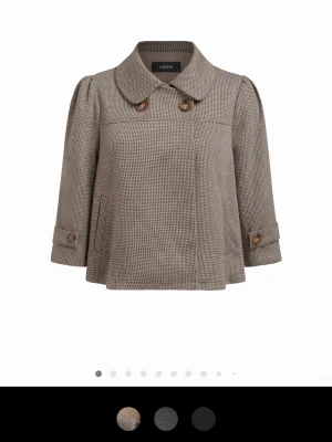 Rutig kort rock från Cider - Kort rock i brunt och beige rutmönster från Cider. Modellen har bred krage, två stora knappar framtill och 3/4-ärmar med knappdetaljer vid ärmslut. Rocken har en lös, A-linjeformad siluett och är tillverkad i ett mjukt tyg med klassisk retrovibe.