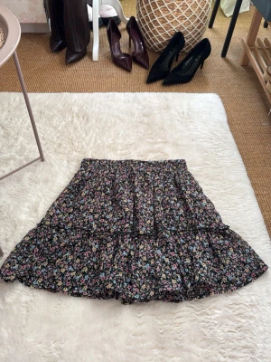 Blommig volangkjol i svart och pastell - Supersöt kort kjol från new yorker i storlek S med blommigt mönster i pastellfärger på svart botten. Super söt för sommriga dagar! 