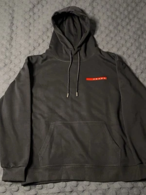 Svart hoodie från Prada  - Snygg svart hoodie från Prada med klassisk röd logga på bröstet. Hoodien har huva med dragsko och en stor magficka framtill. Tillverkad i mjukt material som känns skönt mot huden. Perfekt för dig som gillar stilrena och exklusiva plagg. STORLEK M, PASSAR S-M. HELT NY ALDRIG ANVÄND.