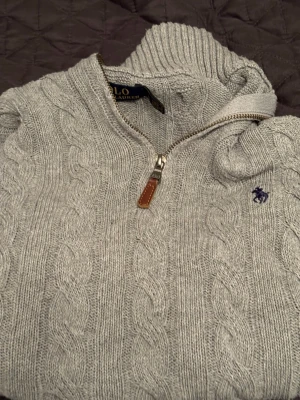 Grå Polo Ralph Lauren stickad i storlek S - Snygg grå kabelstickad tröja från Polo Ralph Lauren med halv dragkedja och hög krage. Tröjan har ribbade muddar och det klassiska broderade logotypen på bröstet. Perfekt för lager på lager och en stilren vardagslook.