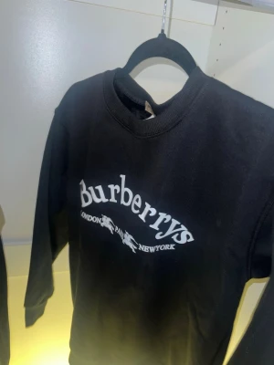 Svart långärmad topp från Burberrys - Säljer den eftersom den var för liten för mig! Svart långärmad topp från Burberrys med vit logga och texten 'London Paris New York' framtill. Klassisk rund halsringning och enkel, stilren design. Perfekt för dig som gillar exklusiva märken och minimalistisk stil.