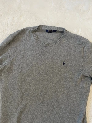 Grå stickad tröja från Polo Ralph Lauren - Klassisk grå stickad tröja från Polo Ralph Lauren med rund halsringning och diskret broderad logga på bröstet. Tröjan har långa ärmar och ribbade muddar vid ärmslut och nederkant. Perfekt till lager på lager och enkel att matcha.