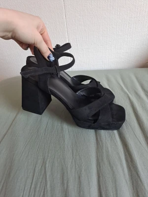 Svarta chunky klackar - Säljer ett par svarta chunky klackar från XIT med bred klack, korsade remmar och fyrkantig tå. Skorna har justerbar ankelrem med spänne och är gjorda i mockaimitation. Haft på mig dessa tre gånger, men de har fått lite synliga märken på klacken då jag hade de på fest