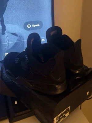 Svarta Air Jordan 4 Retro sneakers - Säljer ett par helsvarta Air Jordan 4 Retro sneakers med klassisk meshpanel på sidan och detaljer i mocka. Skorna har en hög siluett och ikonisk Jumpman-logga på plösen. Perfekta för dig som gillar streetwear och vill ha en stilren look.