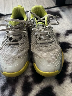 Gråa träningsskor från Craft - Snygga gråa träningsskor från Craft med gula detaljer och svart sula. Skorna har mesh-ovandel för bra ventilation, robust snörning och vadderad insida i gult. Perfekta för inomhussport eller gym. Sportig design med coola svarta prickar på sidan.