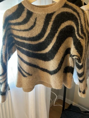 Beige och svart zebramönstrad tröja - Stickad tröja i beige med svart zebramönster. Tröjan har rund halsringning, lång ärm och är gjord i ett fluffigt, mjukt material som ger en cozy känsla. Perfekt för dig som vill sticka ut med ett djurmönster i garderoben.