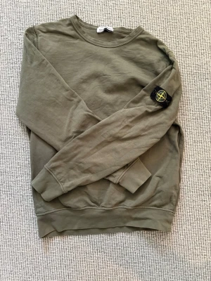 Olivgrön sweatshirt från Stone Island - Snygg olivgrön sweatshirt från Stone Island med klassisk rund halsringning och ribbade muddar.med den iconoska loggan på vänster arm. På höger arm har den dock ett litet litet hål som inte är synligt när den är på. Kan gå ner i pris