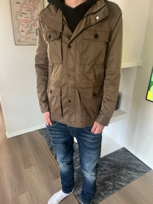  vindjacka - Snygg brun vindjacka med fyra stora fickor framtill. Jackan har knapp- och dragkedjestängning. Tillverkad i ett lätt och vindtätt material, perfekt för blåsiga dagar. Passar dig som gillar en enkel men stilren look.