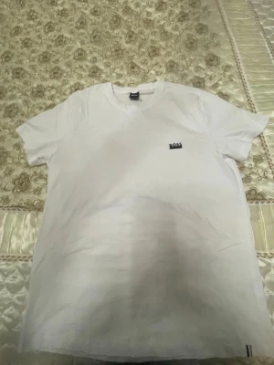 Vit t-shirt från BOSS - Säljer en klassisk vit t-shirt från BOSS med liten logga på bröstet. T-shirten har rund hals och korta ärmar, perfekt för en clean och stilren look. Tillverkad i mjuk bomull för maximal komfort.