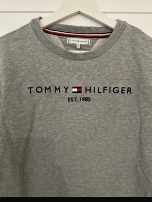 Grå sweatshirt från Tommy Hilfiger - Snygg grå sweatshirt från Tommy Hilfiger med broderad logga och text 'EST. 1985' på bröstet. Tröjan har rund halsringning och ribbade muddar. Tillverkad i mjukt bomullsmaterial och har klassiska Hilfiger-detaljer i rött, vitt och blått vid kragen.