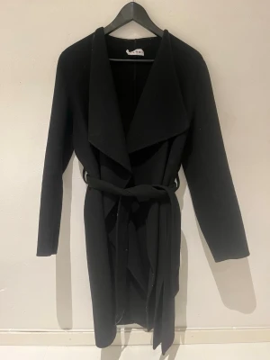 Svart kappa med skärp från Wera - Stilren svart kappa från Wera med bred krage och knytband i midjan. Kappan har en klassisk, rak passform och är tillverkad i ett mjukt material som passar perfekt för kyligare dagar. Enkel att matcha med olika outfits och ger en snygg siluett.