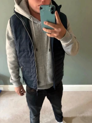 Mörkblå dunväst med huva - Snygg mörkblå dunväst med dragkedja och huva. Västen har två fickor med dragkedja och quiltad design. Perfekt att slänga över en hoodie för en avslappnad och trendig look. Tar även imot byten