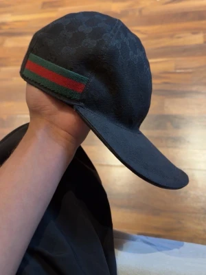 Svart keps från Gucci med GG-mönster - Snygg svart keps från Gucci med klassiskt GG-mönster i tyget och en grön-röd rand på sidan. Justerbar rem bak i läder och böjd skärm. Perfekt accessoar för dig som vill ha en exklusiv och trendig look.
