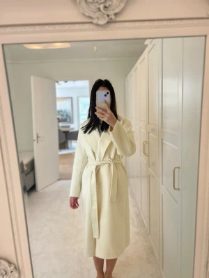 Ny vit Krämvit lång superfin kappa med knytskärp ullkappa från Uniqlo - Perfekt inför vår sommar höst.  Stilren krämvit kappa i lång modell med breda slag och knytskärp i midjan. Kappan har en rak siluett och är tillverkad i ett mjukt material som ger en lyxig känsla. Perfekt för dig som vill ha en clean och elegant look. Nypris 1200kr ny. Passar även xs-s