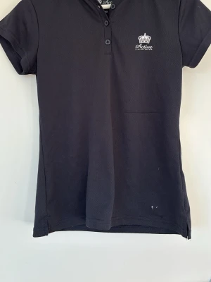 Svart ridtopp från Active Riding Wear - Snygg svart ridtopp från Active Riding Wear med korta ärmar och krage. Toppen har knappar framtill och ett diskret tryck med logga och krona på bröstet. Perfekt för dig som gillar stilren och sportig look. Finns tecken på användning men är i ett gott skick. 
