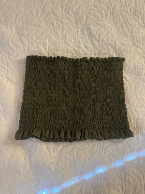 Olivgrön smockad tubtopp med volang - Trendig olivgrön tubtopp med smockad resår och volangkant upptill och nedtill. Perfekt att styla till jeans eller kjol för en cool och avslappnad look. Materialet känns mjukt och följsamt mot huden.