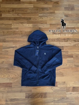 Mörkblå hoodie från Ralph Lauren - 🖤 Ralph Lauren Zip Hoodie – Marinblå | S 🖤  Stilren Ralph Lauren zip hoodie i marinblå, perfekt för vardag och avslappnade tillfällen. Tillverkad i mjuk bomull som ger komfort hela dagen. Lång ärm, ribbade fållar och ärmslut samt vit broderad Ralph Lauren-logga på bröstet. Dragkedja framtill och justerbar huva med snören för anpassad passform. Taggen klippt, nyskick. Ett tidlöst och mångsidigt plagg som passar alla tillfällen.