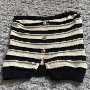 Randig kortkjol med guldknappar - Säljer en svart och vit randig kortkjol med tre dekorativa guldknappar framtill. Kjolen har en ribbad kant nedtill och är tillverkad i ett stickat material som ger en skön och stretchig passform. Perfekt för dig som gillar en trendig och ungdomlig stil.