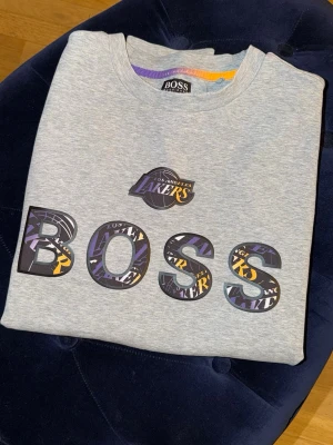 Grå Lakers x BOSS hoodie - Snygg grå hoodie från BOSS i samarbete med Los Angeles Lakers och NBA. Stor BOSS-logga framtill med Lakers-tryck i bokstäverna och patch nertill. Mjuk bomullskänsla och klassisk passform, perfekt för dig som gillar basket och streetwear.