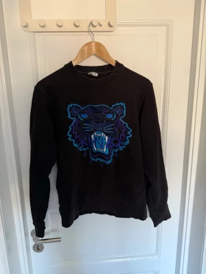 Svart Kenzo sweatshirt med tigertryck - Svart sweatshirt från Kenzo med tigertryck i blått och lila på bröstet. Tröjan har rund halsringning, ribbade muddar vid ärmslut. Vid fler frågor eller intresse för fler bilder så är det bara att höra av sig🤠
