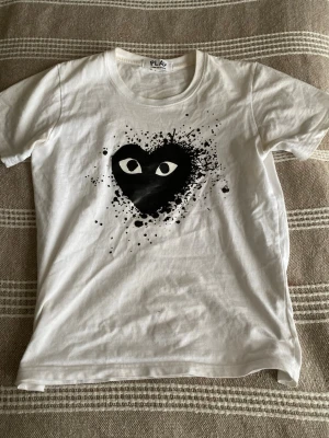 Vit Comme des Garçons PLAY t-shirt - Vit t-shirt från Comme des Garçons PLAY med det ikoniska svarta hjärtat med ögon tryckt på bröstet. T-shirten har rund halsringning och korta ärmar. Tillverkad i mjuk bomull som känns skön mot huden. Perfekt för dig som gillar streetwear och grafiska prints.