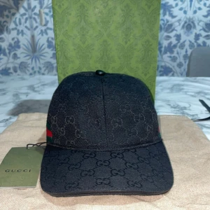 Svart Gucci keps med GG-mönster - Snygg svart keps från Gucci med klassiskt GG-monogram över hela kepsen. På sidan finns en grön och röd rand som ger den en ikonisk touch. Kepsen har böjd skärm och justerbar rem bak. Tillverkad i ett slitstarkt tyg som känns exklusivt. Pris kan diskuteras vid snabb affär, skickar samma dag🤩 (Notera att den är äkta) och kvaliten är på top