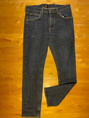 Mörkblå jeans från Lee - Säljer ett par mörkblå jeans från Lee med klassisk femficksdesign och raka ben. Jeansen har guldfärgade sömmar och Lee-logga på bakfickan. Materialet är denim i bomull, perfekt för en avslappnad och stilren look.