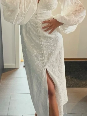 Adoore klänning  - Elegant vit långklänning med broderade blommor, puffiga långärmar och knappar framtill. Klänningen har en v-ringad urringning och en slits framtill för extra stil. Perfekt för sommarens speciella tillfällen.