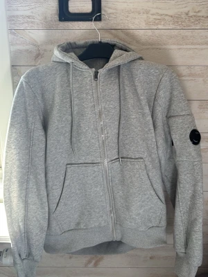 Grå zip hoodie från C.P. Company - En nästan helt ny Snygg grå hoodie med dragkedja från C.P. Company. Klassisk modell med huva och dragsnören, två stora fickor fram och den ikoniska linsen på vänster ärm. Tillverkad i mjuk bomullsblandning för skön komfort och stilren look. Pris kan diskuteras!