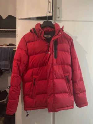 Röd Dressmann Pufferjacka - Snygg röd pufferjacka med huva och svarta detaljer. Jackan har dragkedja framtill, två sidofickor med dragkedja och en bröstficka med svart dragkedja. Perfekt för kalla vinterdagar tack vare den tjocka vadderingen och den höga kragen.