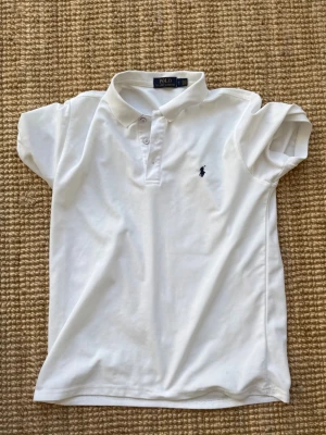 Vit piké från Polo Ralph Lauren - Klassisk vit pikétröja från Polo Ralph Lauren med korta ärmar och liten broderad logga på bröstet. Tröjan har krage och knappar. Den är L men passformen är M