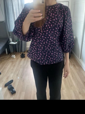 Mörkblå blus med rosa blommor - Supersöt mörkblå blus med små rosa blommor och puffiga långärmar. Blusen har rund halsringning och en lätt figursydd passform. Perfekt för dig som gillar en feminin och trendig stil. Materialet känns lätt och luftigt.