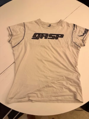 Vit GASP t-shirt med tryck - Säljer en vit t-shirt från GASP med svart broderad logga på bröstet och coola kontrastsömmar. T-shirten har rund halsringning och korta ärmar. Materialet känns mjukt och skönt, perfekt för en avslappnad stil.