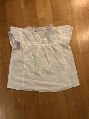 Vit blus med volangärm från Kappahl - Söt vit blus från Kappahl med korta volangärmar och liten v-ringning framtill. Blusen har en avslappnad passform och är gjord i mjuk bomull, perfekt för varma dagar. Enkel och stilren design som är lätt att matcha. Aldrig använd och säljer då jag upplever att den har blivit för kort. 