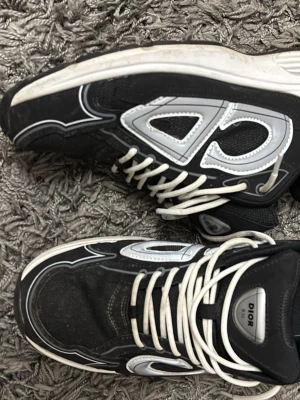 Svarta  sneakers - Säljer ett par svarta  sneakers med vita och grå detaljer samt stora reflekterande på sidorna. Skorna har vita snören, tjock sula och meshpartier för extra komfort. Perfekta för dig som vill ha en statement sneaker med lyxig känsla.