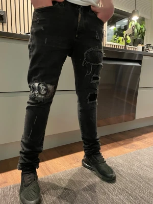 Svarta slitna jeans med tryck - Säljer ett par svarta jeans med slitningar och tryck på vänster lår. Jeansen har flera hål och patchade detaljer på framsidan, vilket ger en riktigt edgy och trendig look. Perfekta för dig som vill sticka ut med din stil.