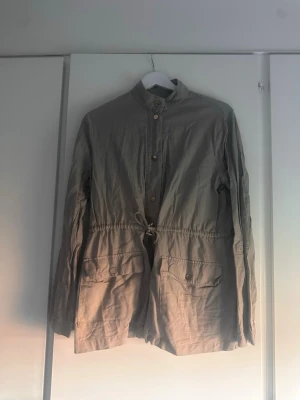 Beige parkajacka med knappar - Stilren army grön parkajacka med hög krage, knappar framtill och dragsko i midjan. Jackan har stora fickor  och långa ärmar som man kan vika upp. Perfekt för dig som gillar en enkel men snygg look.