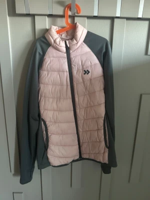 Ljusrosa quiltad jacka från 157 - Snygg ljusrosa quiltad jacka från 157 med grå ärmar och detaljer. Jackan har dragkedja framtill, två fickor och en sportig look. Perfekt för dig som gillar en stilren och bekväm jacka till vardags. Tar emot förslag på priset