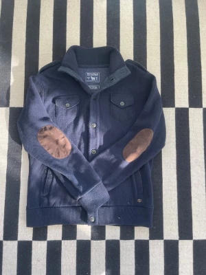 Marinblå cardigan från Woolrich - Snygg marinblå cardigan från Woolrich med bruna mockadetaljer på armbågarna. Tröjan har två bröstfickor med knapp, ribbade muddar och hög krage. Tillverkad i en mjuk ull- och bomullsblandning som känns både lyxig och bekväm.