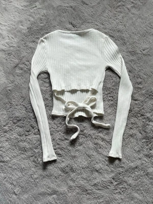 Vit ribbad topp med knyt från FB Sister - Trendig vit ribbad topp från FB Sister med långa ärmar och snygg knytning i midjan. Toppen är croppad och har en tight passform som framhäver figuren. Perfekt att styla med höga jeans eller kjol för en cool look.