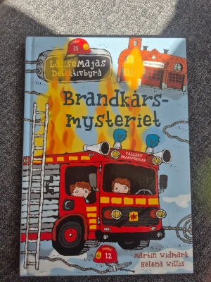 Brandkårsmysteriet LasseMajas Detektivbyrå - En färgglad och illustrerad barnbok i serien LasseMajas Detektivbyrå, skriven av Martin Widmark och illustrerad av Helena Willis. Omslaget visar en röd brandbil, eldslågor och Valleby brandstation. Boken har hårda pärmar och är på svenska.