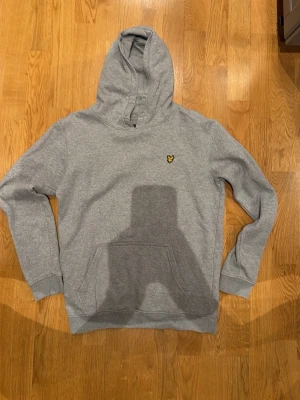 Grå hoodie från Lyle & Scott Junior - Snygg grå hoodie från Lyle & Scott Junior med klassisk gul logga på bröstet. Hoodien har en stor huva och en praktisk magficka. Tillverkad i mjukt bomullsmaterial som är skönt att ha på sig. Perfekt för en avslappnad och stilren look.