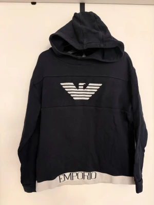 Mörkblå hoodie från Emporio Armani - Snygg mörkblå hoodie från Emporio Armani med stor logga i vitt framtill och texten 'EMPORIO' och 'ARMANI' på den vita mudden nedtill. Klassisk huva och lång ärm. Tillverkad i mjukt bomullsmaterial som känns skönt mot huden.