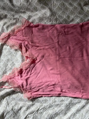 Rosa ribbad linnetopp med spets - Söt rosa linnetopp med tunna axelband och ribbad struktur. Toppen har spetsdetaljer längs urringningen och axelbanden för en romantisk vibe. Perfekt att styla lager på lager eller bära som den är under varma dagar. Justerbara axelband