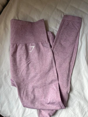 Lila/rosa seamless träningsbyxor från Gymshark - Säljer ett par lila/rosa seamless leggings från Gymshark med hög midja och ribbad linning. De har diskret strukturmönster längs benen och en liten vit logga på höften. Materialet är stretchigt och mjukt. Vet inte storleken men skulle säga s/m 