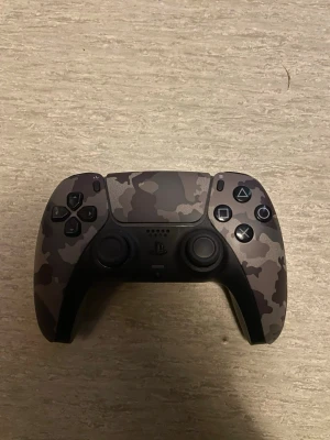 PS5 DualSense Camo handkontroll - Snygg PlayStation 5 DualSense handkontroll i grått camomönster. Ergonomisk design med klassiska PlayStation-knappar och touchpad. Tillverkad i slitstark plast och passar perfekt för gaming på PS5.
