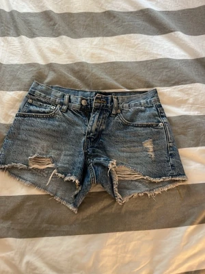 Blå jeansshorts med slitningar - Säljer ett par blå jeansshorts från 157 med råa kanter och slitna detaljer framtill. Klassisk femficksmodell med knapp och dragkedja. Perfekta till sommaren för en avslappnad look.