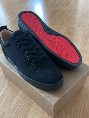 Christian Louboutin skor - Snygga svarta sneakers från Christian Louboutin med klassisk röd sula och svarta skosnören. Skorna är tillverkade i mocka och har en låg profil. Kommer med originalkartong, dustbag och certifikat.