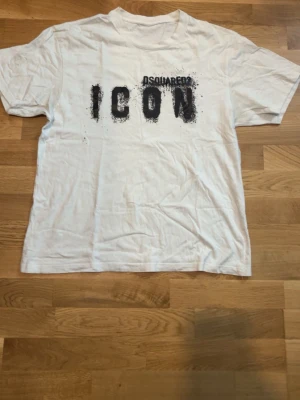 Vit Dsquared2 ICON t-shirt - Säljer en vit t-shirt från Dsquared2 med svart tryck där det står 'ICON' på bröstet. T-shirten har korta ärmar och en klassisk passform. Perfekt för dig som gillar streetwear och vill ha en statement-piece i garderoben.