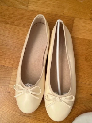 Krämvita ballerinaskor med rosett - Snygga ballerinaskor i krämvitt med rund tå och söt rosettdetalj framtill. Skorna har en slät ovandel i konstläder och en mjuk innersula för extra komfort. Yttersulan är i gummi för bra grepp. Perfekta till jeans eller kjol för en clean look.helt nya 